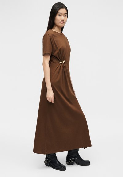 Γυναίκα που φοράει καφέ μακρυμάνικο φόρεμα με κοντά μανίκια και σούρες στη μέση, διακοσμημένο με χρυσό καρφίτσα σε σχήμα φτερού, σε συνδυασμό με μαύρες μπότες αστραγάλου με αλυσίδες.