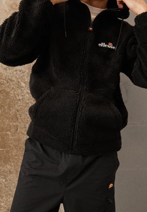 Personne portant une veste polaire noire zippée et un pantalon noir assorti, tous deux avec le logo Ellesse, ajustant le col de la veste.