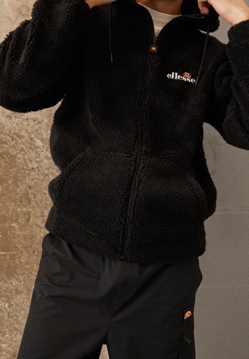 Personne portant une veste polaire noire zippée et un pantalon noir assorti, tous deux avec le logo Ellesse, ajustant le col de la veste.
