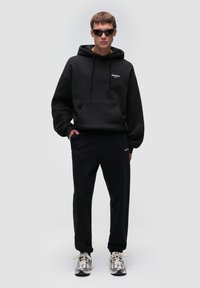 Sudadera negra con bolsillo frontal y cordones, combinada con pantalones de chándal azul marino oscuros. El conjunto incluye zapatillas deportivas gris claro y gafas de sol negras.