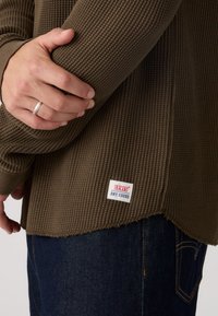 Καφέ μπεζ πουλόβερ με μακριά μανίκια και ταμπέλα που γράφει "LEVI'S DRY GOODS", συνδυασμένο με σκούρο μπλε τζιν τζιν. Έμφαση στην υφή και στη στρώση των υλικών.