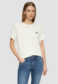 Witte katoenen T-shirt met een klein blauw hartontwerp op de borst, gecombineerd met blauwe denim jeans. Het T-shirt heeft korte mouwen en een ronde hals.