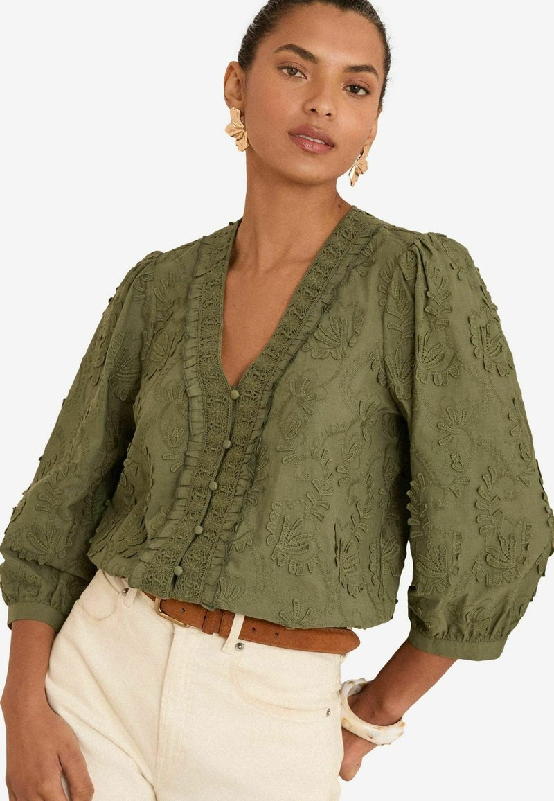 Blouse verte brodée avec un décolleté en V, des manches bouffantes et des boutons décoratifs. Présente des motifs floraux texturés et une finition froncée.
