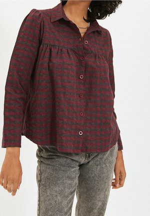 Frau trägt eine rot-grau karierte Bluse mit Knöpfen und gerafftem Brustdetail sowie grau gewaschene Jeans, steht vor einem einfarbigen Hintergrund.