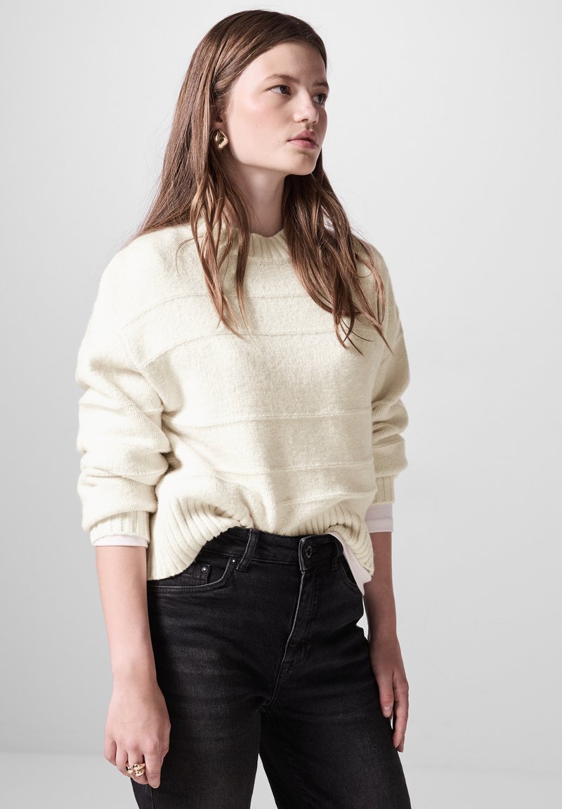 Maglione lavorato a maglia color crema con texture a righe orizzontali e orlo a coste, abbinato a jeans neri a vita alta.
