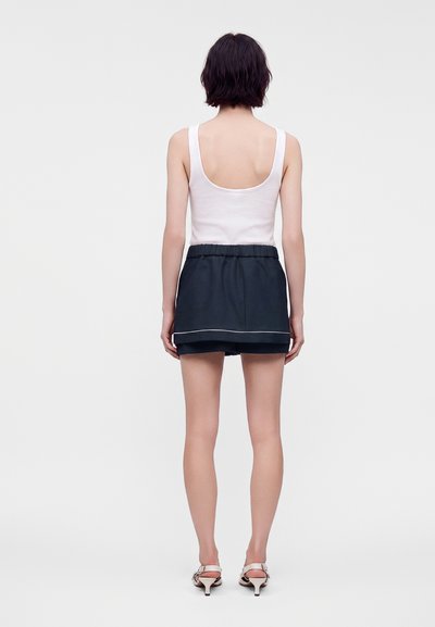 Femme aux cheveux courts et foncés portant un haut blanc sans manches, un short noir et des sandales blanches à talons, debout, dos tourné vers l'avant.