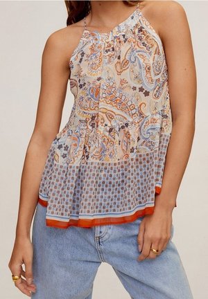 Kvinde iført en ærmeløs, mønstret bluse med orange, blå og brune paisley- og geometriske designs, kombineret med lyseblå jeans.