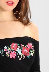 Schwarzes Off-Shoulder-Top mit buntem Blumenstickereien in Rot, Pink und Blau. Glattes Baumwollmaterial, taillierter Schnitt und keine sichtbaren Hardware.