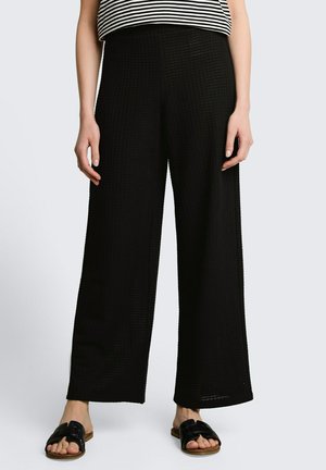 Kobieta w czarnych, teksturowanych spodniach z szerokimi nogawkami, czarnych klapkach typu slide i czarno-białej paskowanej bluzce, stojąca na jednolitym tle.