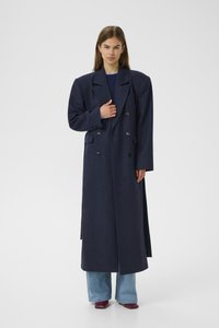 Navy wool blend double-breasted coat with pointed lapels, side pockets, and straight silhouette; paired with light blue jeans and red shoes.