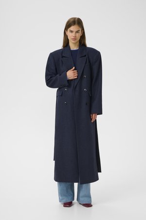 Navy wool blend double-breasted coat with pointed lapels, side pockets, and straight silhouette; paired with light blue jeans and red shoes.
