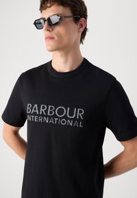 T-shirt de algodão preto com o logo "BARBOUR INTERNATIONAL" em material reflexivo. O decote redondo tem uma textura canelada.