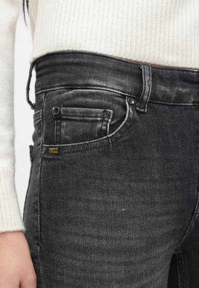 Λεπτά μαύρα τζιν με ξεθωριασμένη υφή, διαθέτουν μπροστινές και πίσω τσέπες, κλείσιμο με κουμπί στο επάνω μέρος και μια μικρή κίτρινη ετικέτα στην τσέπη.