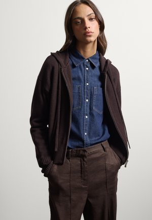 Joven mujer con una sudadera marrón con cremallera sobre una camisa vaquera azul con botones y pantalones marrones, con las manos en los bolsillos, sobre un fondo liso.