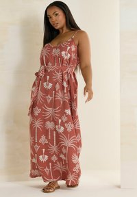 Maximeklänning i terrakotta med vita palmträd och blommönster. Spaghetti band och knytning i midjan. Platta bruna sandaler synliga.