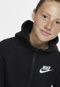 Črna Nike zip-up kapuca z visokim ovratnikom, ki ima belo logo na prsih in gladko teksturo tkanine.