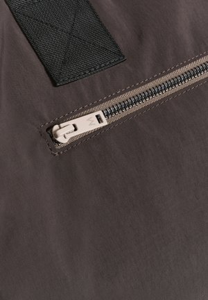 Tmavohnedá tkanina zipsová kapsa s textúrovaným povrchom, s béžovým zipsom a čiernym nylonovým popruhom, zdôrazňujúca čisté línie a minimalistický dizajn.