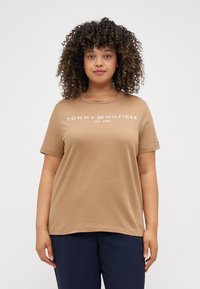 Šviesiai ruda medvilninė marškinėliai su trumpomis rankovėmis, apvaliu iškirpimu ir balta "TOMMY HILFIGER EST. 1985" spauda per krūtinę.