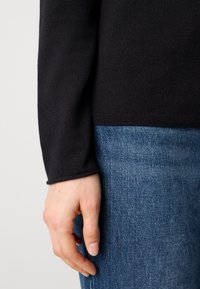 Main gauche détendue à côté d'un jean en denim bleu et d'un pull noir à manches longues visible du poignet à la taille.