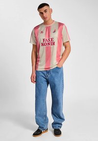 Ljusbeige och rosa randig t-shirt med logotyp, kombinerad med blå baggy jeans och svarta skor. T-shirten har en rund halsringning och korta ärmar.