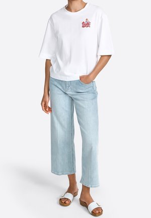 Persoon draagt een wit oversized T-shirt met een kleine kleurrijke afbeelding op de borst, lichtblauwe wijde cropped jeans en witte sleehakken.