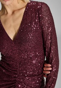 Robe en sequins bordeaux avec un décolleté en V profond, des manches longues et des détails froncés à la taille. Texture scintillante avec des sequins réfléchissants.