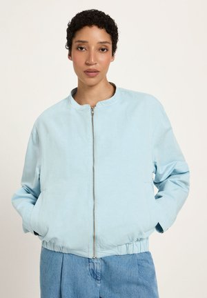 Woman with short curly hair wearing a light blue zip-up bomber jacket and blue jeans, hands in jacket pockets, neutral background.