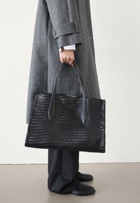 Sac fourre-tout tissé noir avec des poignées plates, fabriqué en matériau texturé. Présente une forme rectangulaire et des accents lisses.