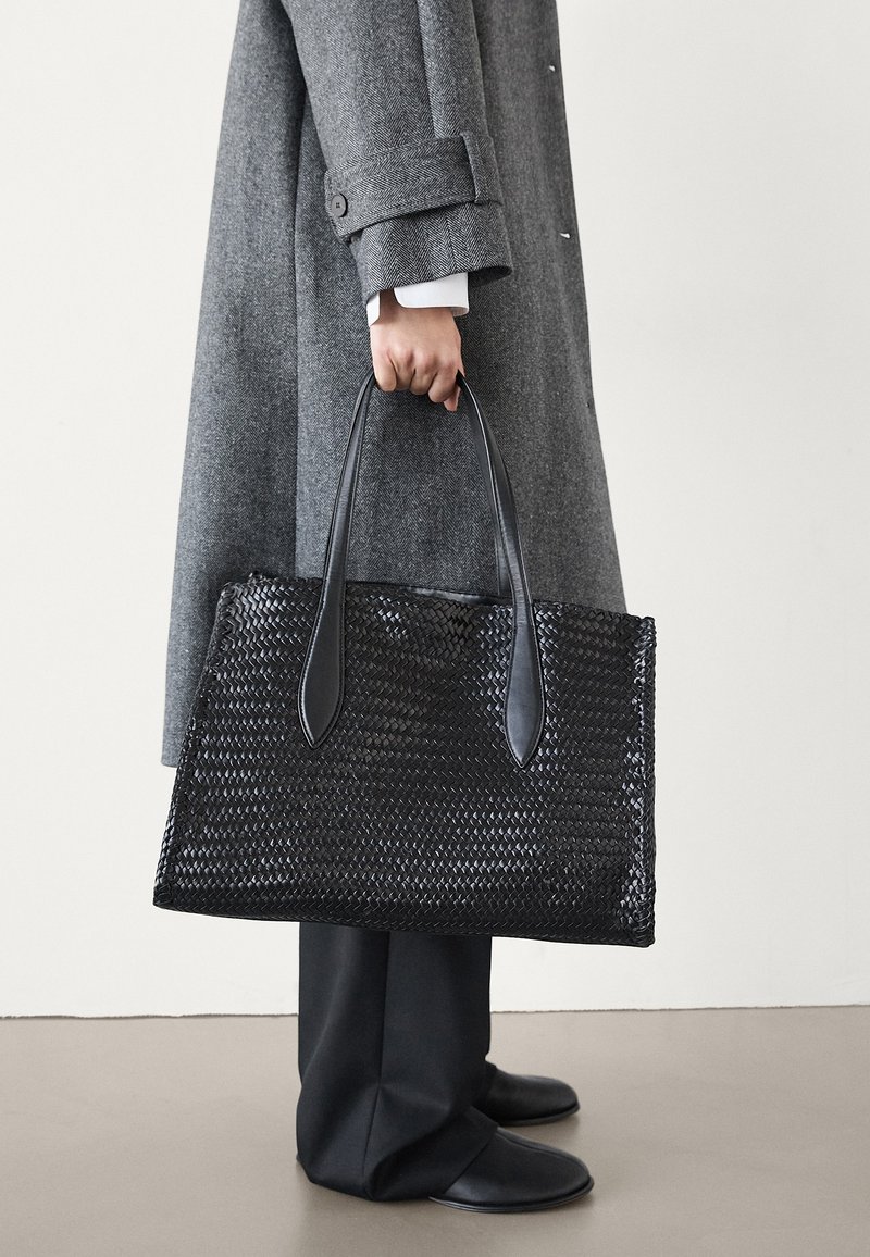 Sac fourre-tout tissé noir avec des poignées plates, fabriqué en matériau texturé. Présente une forme rectangulaire et des accents lisses.