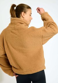Pullover in pile marrone con colletto alto, vestibilità ampia, superficie testurizzata e spalle cadenti, abbinato a leggings neri.