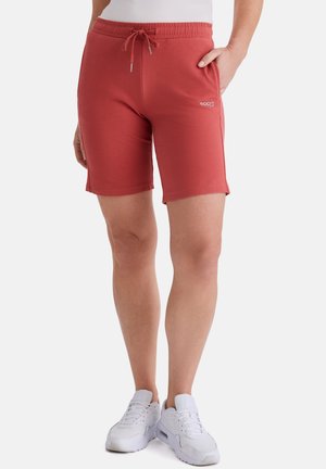 Person trägt kniehohe rote Shorts mit Kordelzug und Seitentaschen sowie weiße Sportschuhe, steht mit einer Hand in der Tasche.