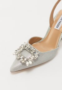 Sølvglitrende høyhælte sko med spiss tå, slingback-design og en stor rhinsteinbesatt spenne detalj foran.