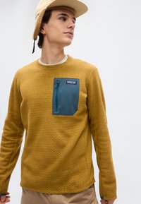 Νεαρός άνδρας που φοράει μουσταρδί κίτρινο υφασμάτινο πουλόβερ με μπλε τσέπη με φερμουάρ και μπεζ καπέλο, κοιτάζοντας ελαφρώς προς το πλάι σε απλό φόντο.