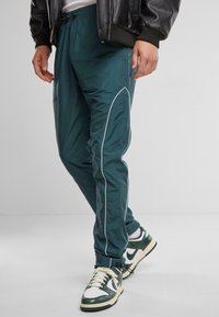 Pantalones deportivos de nailon en color verde azulado con ribetes blancos a los lados, cintura elástica y un ajuste tapered, combinados con zapatillas verdes y blancas.
