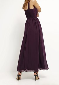 Kvinna i en ärmlös, golvlång plommonlila klänning med smockad midja, stående med armarna i kors, iförd svarta högklackade sandaler.