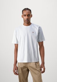 Weißes Baumwoll-T-Shirt mit kurzen Ärmeln, Rundhalsausschnitt und kleinem roten Dreieckslogo auf der linken Brust; kombiniert mit hellbeigen Hosen.