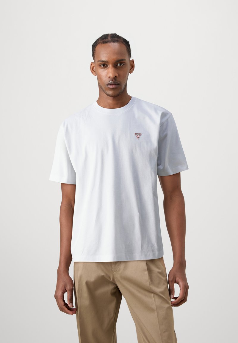 Weißes Baumwoll-T-Shirt mit kurzen Ärmeln, Rundhalsausschnitt und kleinem roten Dreieckslogo auf der linken Brust; kombiniert mit hellbeigen Hosen.