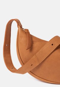 Tasche aus braunem Leder mit rundem Design, strukturiertem Material und abnehmbarem Tragegurt. Ausgestattet mit einem Reißverschluss und einem kleinen Lederanhänger als Akzent.