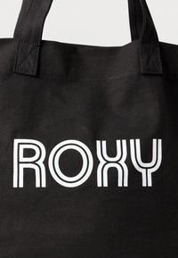 Melnā soma ar rokturi, kas izgatavota no izturīgas auduma, uz kuras redzams balts uzraksts "ROXY" izteiksmīgā, modernā fontā ar apļveida elementiem. Rokturi izturīgi šūti.