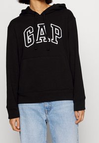 Sweat à capuche noir en coton, avec un logo "GAP" blanc sur le devant, capuche à cordon de serrage et poche kangourou, porté avec un jean bleu.