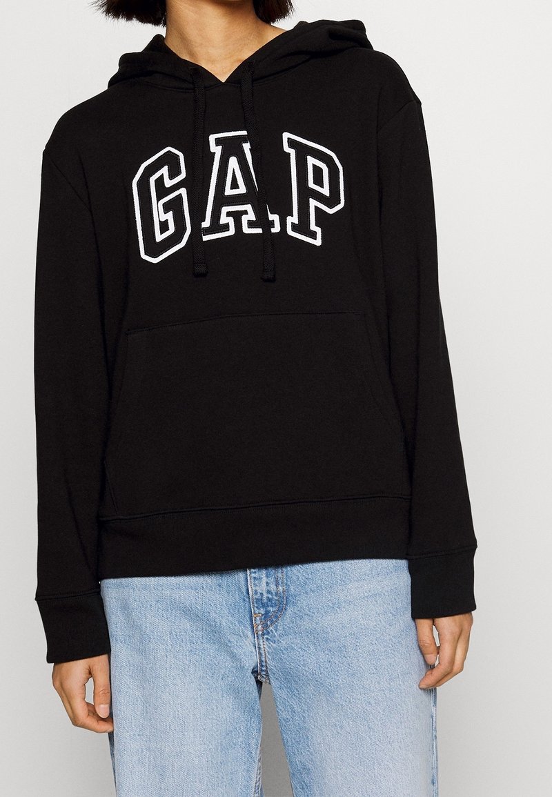 Sweat à capuche noir en coton, avec un logo "GAP" blanc sur le devant, capuche à cordon de serrage et poche kangourou, porté avec un jean bleu.