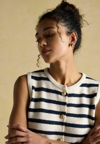 Mouwloze, crème en marineblauwe gestreepte truitje met ronde halslijn en goudkleurige knoopdetails, voorzien van een zachte textuur.