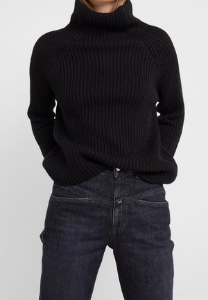 Personne portant un pull à col roulé côtelé noir et un jean bleu foncé, les mains dans les poches, sur un fond uni.