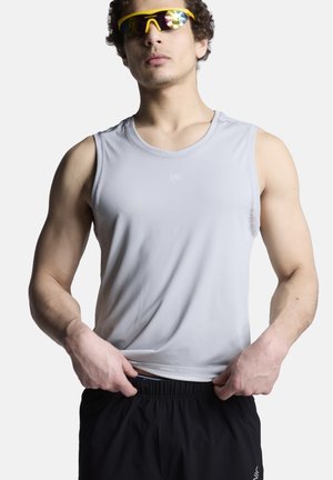 Man draagt lichtgrijs mouwloos sportshirt en zwarte shorts, past de zoom van zijn shirt aan, draagt gele sportzonnebril met reflecterende lenzen.