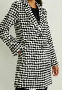Manteau à motif pied-de-poule noir et blanc, doté d'un col cranté, de deux boutons frontaux et de poches latérales, avec une coupe cintrée.