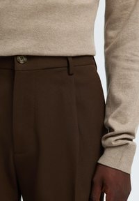 Μπεζ μακρυμάνικη μπλούζα φορεμένη μέσα σε καφέ παντελόνι ψηλόμεσο, φορεμένη από άτομο με σκούρο δέρμα, που φαίνονται η μέση και το δεξί χέρι.