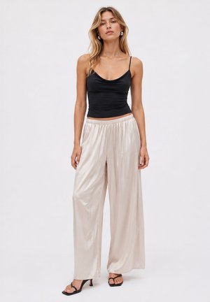Donna in piedi che indossa un top nero a spaghetti, pantaloni larghi in raso beige e sandali neri con tacco su uno sfondo chiaro e semplice.