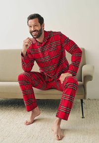 Rød ternet flannel pyjamas med sorte og grønne detaljer; lange ærmer, skjorte med knapper og matchende bukser; siddende på en lys farvet sofa.