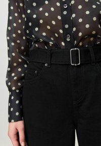 Schwarze, transparente Bluse mit weißen Punkten und Knopfleiste vorne, kombiniert mit hoch taillierter schwarzer Denim-Jeans mit rechteckiger silberner Schnalle am Gürtel.