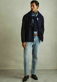 Marinfärgad dubbelknäppt kappa över en ljusblå skjorta, urvuxna blå jeans och mörka loafers. Knytstild marinscarf hängande runt halsen.